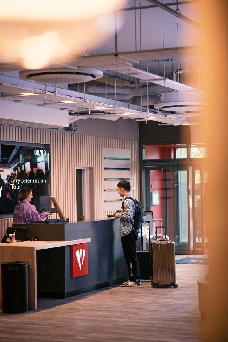 A person stands at a hotel check-in counter with a large suitcase. They're interacting with a receptionist. The interior is modern with a digital display announcing an "Orientation Tour." The space is well-lit with ceiling fixtures.